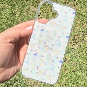 Pode incluir: Uma capa transparente para smartphone com um padrão repetitivo de pequenas personagens de desenhos animados, incluindo ursos e coelhos, intercaladas com motivos florais coloridos. A capa tem um design de anel magnético circular.