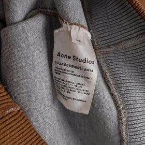 May include: A close-up shot of a grey and brown garment with a label from Acne Studios. The label states "COLLEGE REVERSE AW14" and details the fabric composition, including cotton, wool, and acrylic. Made in China.