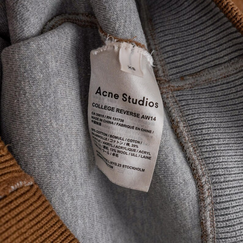 May include: A close-up shot of a grey and brown garment with a label from Acne Studios. The label states "COLLEGE REVERSE AW14" and details the fabric composition, including cotton, wool, and acrylic. Made in China.