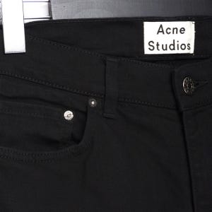 May include: Close-up of black denim jeans with the "Acne Studios" label visible. The jeans have a classic five-pocket design with silver rivets and a button closure. The fabric appears to be a sturdy, dark wash denim.