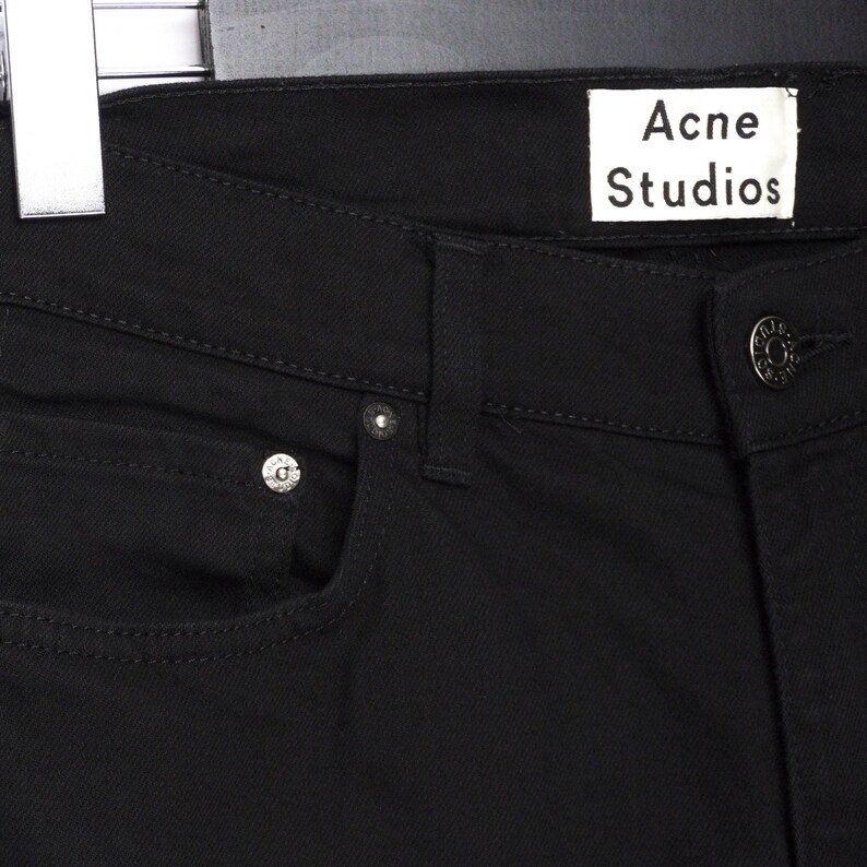 May include: Close-up of black denim jeans with the "Acne Studios" label visible. The jeans have a classic five-pocket design with silver rivets and a button closure. The fabric appears to be a sturdy, dark wash denim.