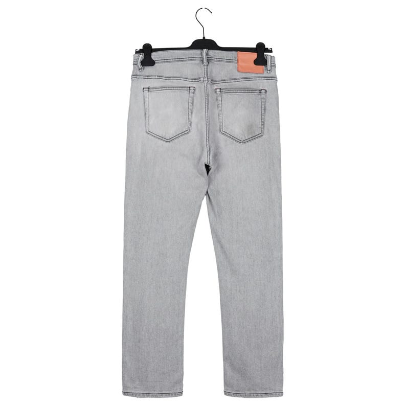 May include: Light gray denim jeans with a classic five-pocket design, hanging on a black hanger. The jeans feature a straight leg cut and a small, rectangular leather patch on the back waistband. The stitching is a darker gray.