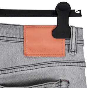 May include: Close-up of light gray denim jeans with a peach-colored leather patch. The patch is embossed with the words "Acne Studios". The jeans are hanging on a black hanger.