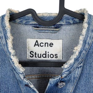 Op de afbeelding: Close-up van een lichtblauwe spijkerjas met een gerafelde kraag. De jas heeft een wit label met de tekst "Acne Studios" in zwart. De jas hangt aan een zwarte hanger.