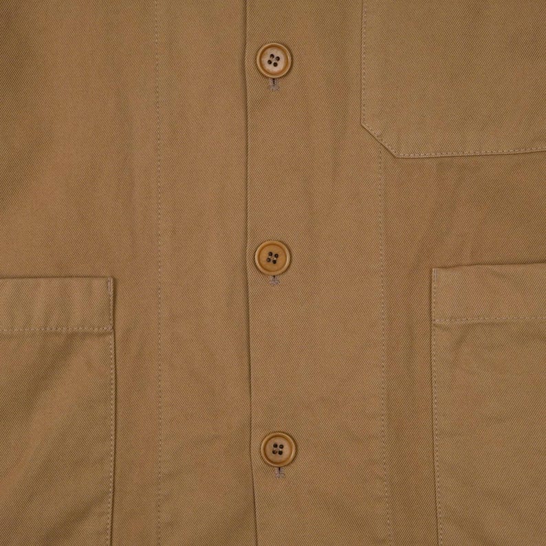 May include: Close-up of a tan-colored jacket with three visible buttons and two front pockets. The jacket has a classic design with a tailored fit, suitable for various occasions. The buttons are a matching tan color.
