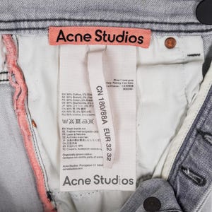 May include: Close-up of light grey denim jeans with the "Acne Studios" label visible. The label includes size information and fabric composition details. The jeans have a button closure and a light pink seam.