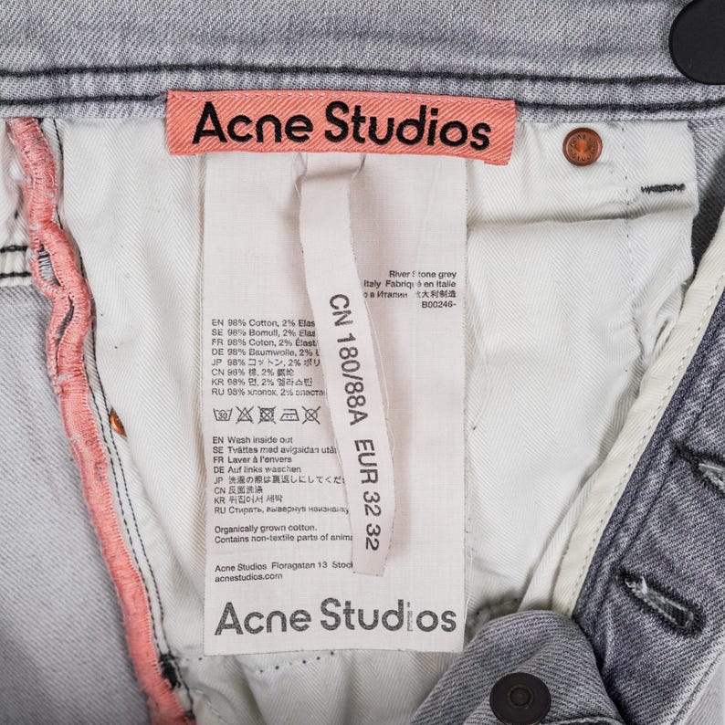 May include: Close-up of light grey denim jeans with the "Acne Studios" label visible. The label includes size information and fabric composition details. The jeans have a button closure and a light pink seam.