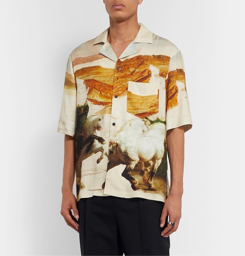 May include: Short-sleeved button-down shirt with a landscape and horse painting design. The shirt features a classic collar, a chest pocket, and a beige, brown, and green color palette. The artwork depicts white horses in a dynamic pose.
