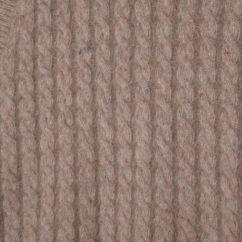 May include: Close-up of a beige cable knit sweater. The knit pattern features vertical columns of intertwined cable stitches. The fabric has a soft texture and subtle flecks of blue and brown throughout, adding visual interest to the design.