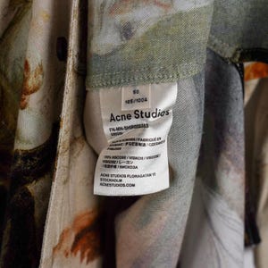 May include: Close-up of a garment tag from Acne Studios. The tag is white with black text, including the brand name, size, and fabric composition. The garment has a floral pattern with shades of green, orange, and beige.
