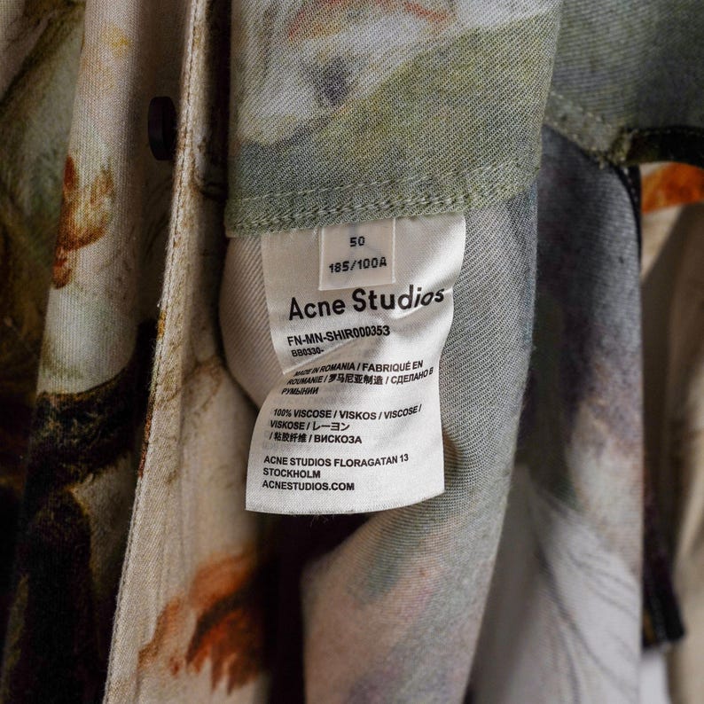 May include: Close-up of a garment tag from Acne Studios. The tag is white with black text, including the brand name, size, and fabric composition. The garment has a floral pattern with shades of green, orange, and beige.