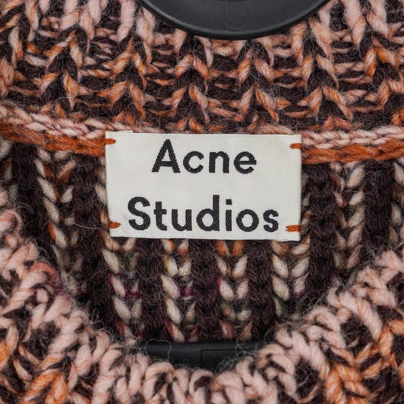 May include: Close-up of a textured knit sweater in shades of brown, orange, and pink. The label reads "Acne Studios" in black text on a white background. The sweater is hanging on a black hanger.