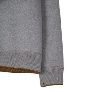 May include: Close-up of a grey sweatshirt sleeve. The ribbed cuffs and hem are a darker grey with a brown accent. The fabric appears to be a soft, comfortable material, suitable for casual wear.