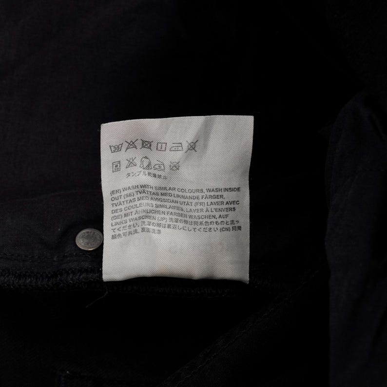 May include: A close-up of a black garment with a white tag attached. The tag features washing instructions and text in multiple languages, including English, French, and German. A silver button is visible.