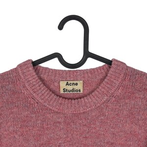May include: A pink crew neck sweater with the "Acne Studios" label. The sweater is hanging on a black hanger. The knitwear has a textured appearance and a round neckline.