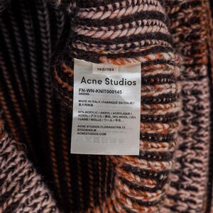 May include: Close-up of a knitted garment with a tag from Acne Studios. The tag displays the brand name, product details, and material composition, including acrylic and wool. The knitwear features a textured pattern in shades of brown, orange, and cream.