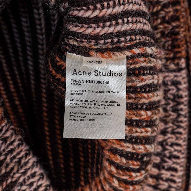 May include: Close-up of a knitted garment with a tag from Acne Studios. The tag displays the brand name, product details, and material composition, including acrylic and wool. The knitwear features a textured pattern in shades of brown, orange, and cream.