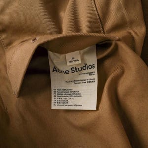 May include: Close-up of a tan-colored garment with a label from Acne Studios. The label includes text in multiple languages, including English, French, and Japanese, detailing the fabric composition as 100% cotton.