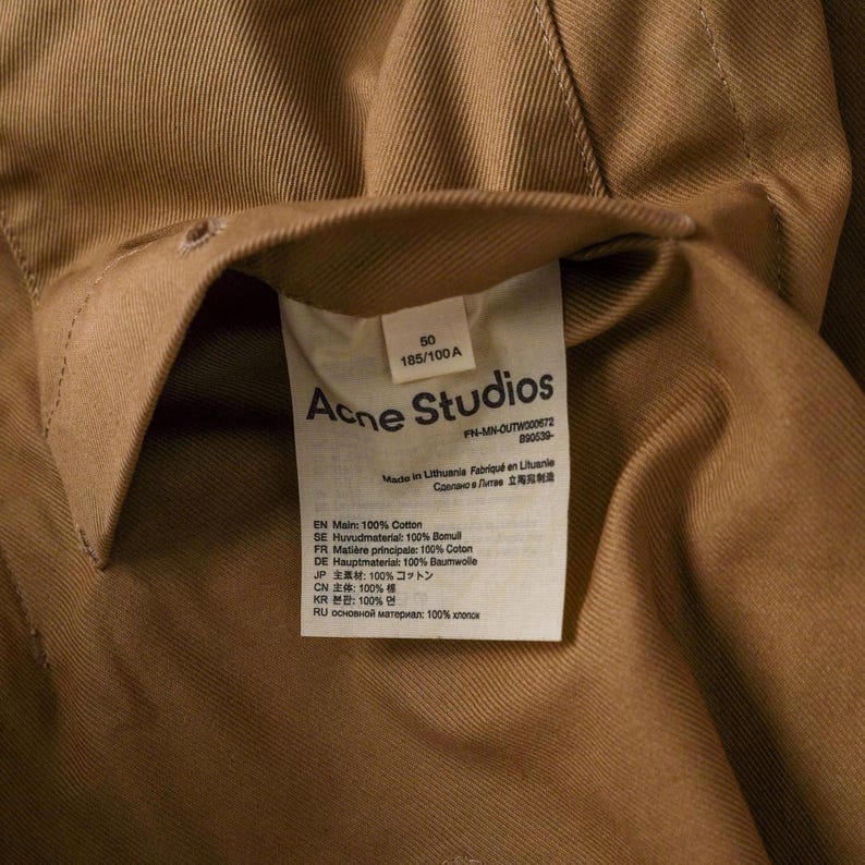 May include: Close-up of a tan-colored garment with a label from Acne Studios. The label includes text in multiple languages, including English, French, and Japanese, detailing the fabric composition as 100% cotton.