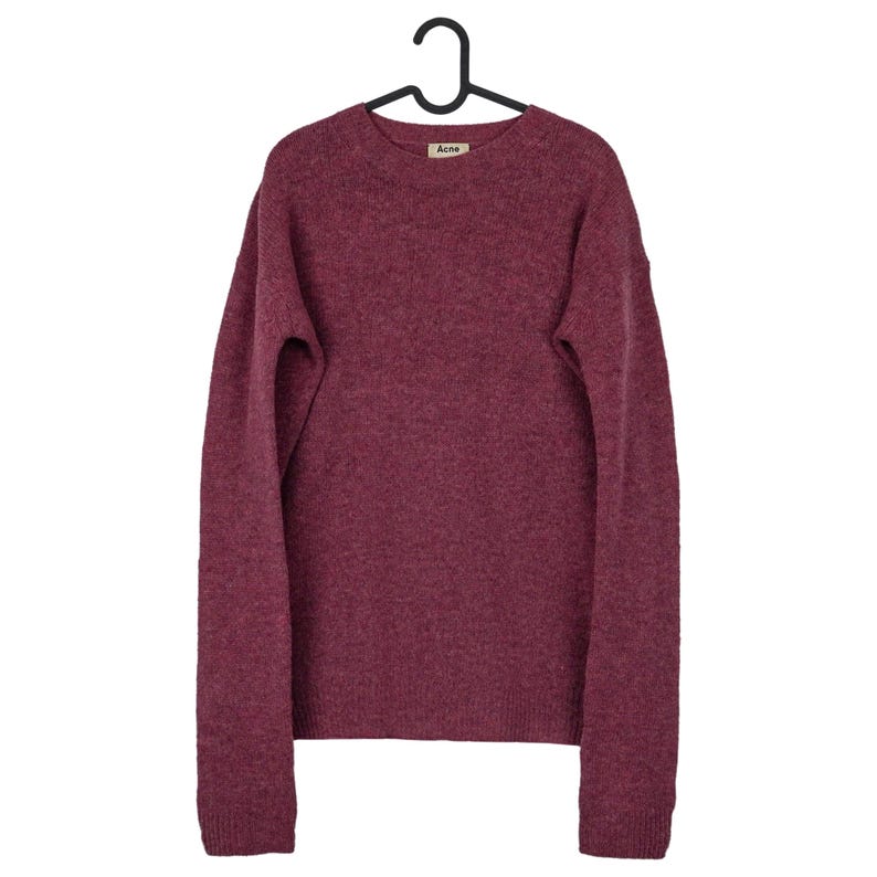 May include: A burgundy crew neck sweater with long sleeves, hanging on a black hanger. The sweater has a small label near the neckline. The fabric appears to be a soft knit.