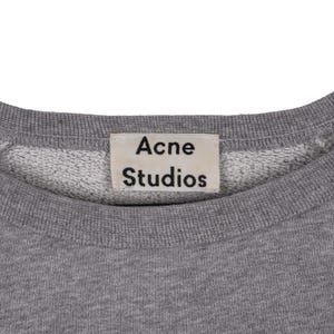 Puede incluir: Primer plano de una sudadera gris de cuello redondo con el nombre de la marca "Acne Studios" impreso en una etiqueta rectangular. El interior del cuello es blanco.