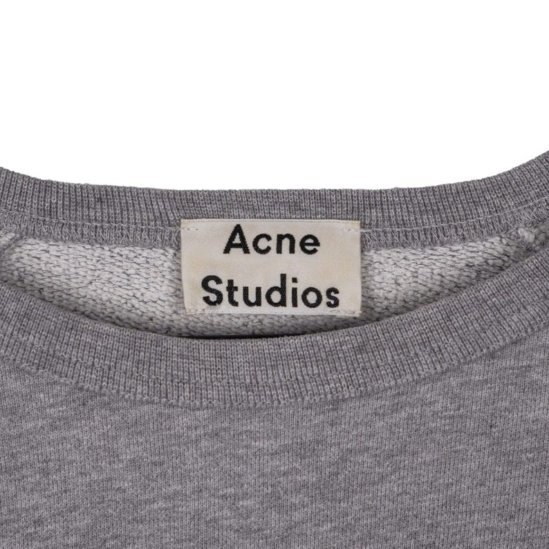 Puede incluir: Primer plano de una sudadera gris de cuello redondo con el nombre de la marca "Acne Studios" impreso en una etiqueta rectangular. El interior del cuello es blanco.