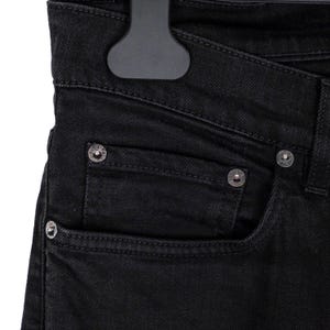 May include: Close-up of black denim jeans with silver rivets and a pocket detail. The jeans are hanging on a black hanger, showcasing the texture and design of the fabric. The focus is on the front pocket and the rivets.