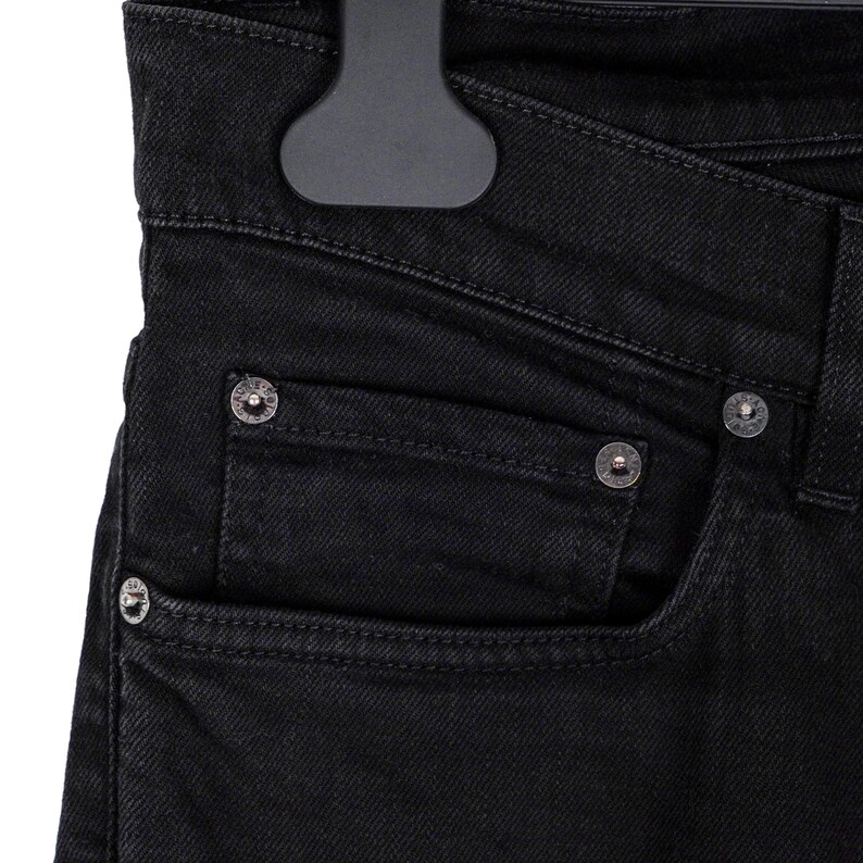 May include: Close-up of black denim jeans with silver rivets and a pocket detail. The jeans are hanging on a black hanger, showcasing the texture and design of the fabric. The focus is on the front pocket and the rivets.