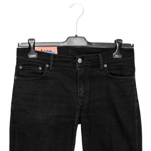 May include: Black denim jeans hanging on a black hanger. The jeans feature a classic five-pocket design with a button and zip closure. A brand tag is visible. The jeans are a solid black colour.