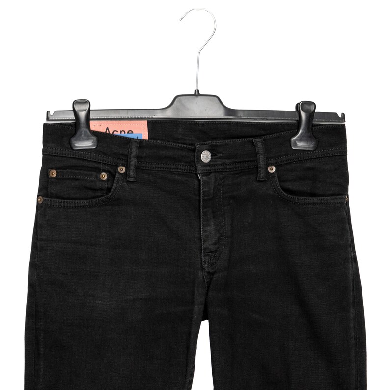 May include: Black denim jeans hanging on a black hanger. The jeans feature a classic five-pocket design with a button and zip closure. A brand tag is visible. The jeans are a solid black colour.