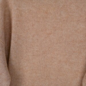 Pu&ograve; includere: Primo piano di un maglione beige. La maglieria ha una consistenza morbida e sfocata e un colore neutro. Le maniche sono visibili sul lato sinistro del fotogramma. Il maglione sembra essere fatto di un materiale caldo e confortevole.