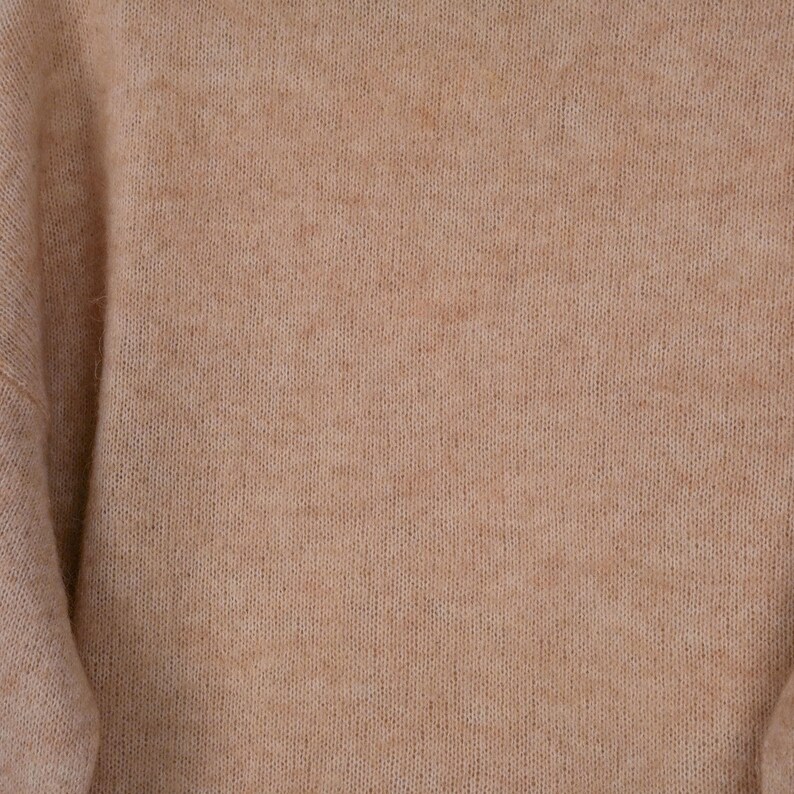 Pu&ograve; includere: Primo piano di un maglione beige. La maglieria ha una consistenza morbida e sfocata e un colore neutro. Le maniche sono visibili sul lato sinistro del fotogramma. Il maglione sembra essere fatto di un materiale caldo e confortevole.