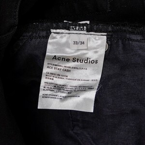May include: Close-up of a black denim garment with a white tag. The tag reads "Acne Studios" and "ACE STAY CASH", with size 33/34. The tag also lists the garment's composition and origin.