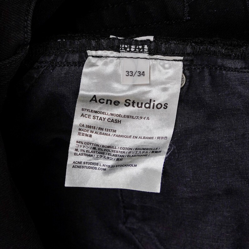 May include: Close-up of a black denim garment with a white tag. The tag reads "Acne Studios" and "ACE STAY CASH", with size 33/34. The tag also lists the garment's composition and origin.
