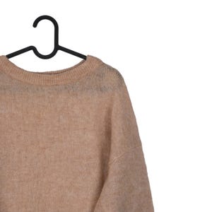 Pu&ograve; includere: Un maglione color beige &egrave; appeso a una gruccia nera su sfondo bianco. Il maglione ha uno scollo tondo e una vestibilit&agrave; ampia e comoda. Il tessuto a maglia appare morbido e leggermente trasparente, suggerendo un design leggero.