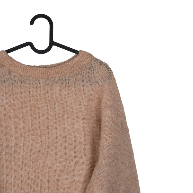 Pu&ograve; includere: Un maglione color beige &egrave; appeso a una gruccia nera su sfondo bianco. Il maglione ha uno scollo tondo e una vestibilit&agrave; ampia e comoda. Il tessuto a maglia appare morbido e leggermente trasparente, suggerendo un design leggero.