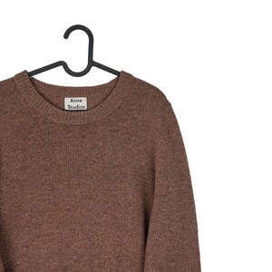 May include: A brown crew neck sweater with the "Acne Studios" label. The sweater is hanging on a black hanger. The sweater is made of a soft, knitted material.