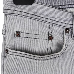 May include: Close-up of light gray denim jeans. The jeans have a classic five-pocket design with copper-colored rivets and black stitching. The fabric has a subtle texture, and the jeans are hanging on a black hanger.