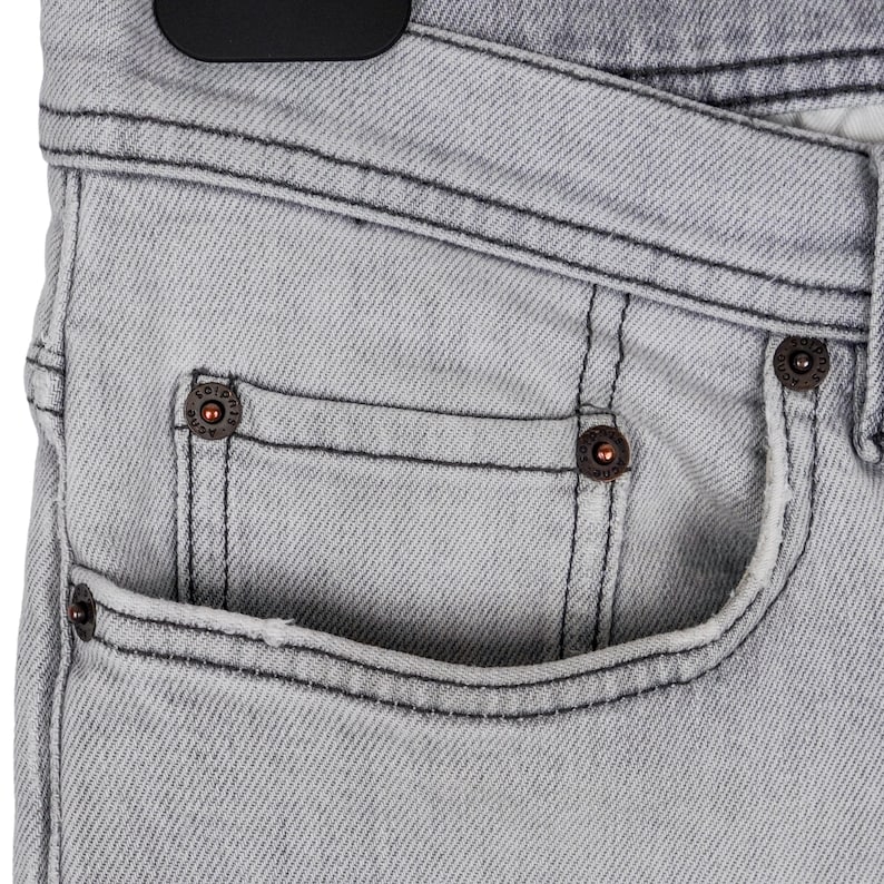 May include: Close-up of light gray denim jeans. The jeans have a classic five-pocket design with copper-colored rivets and black stitching. The fabric has a subtle texture, and the jeans are hanging on a black hanger.
