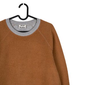 May include: A brown fleece sweatshirt with a grey crew neck and cuffs, hanging on a black hanger. The label "Acne Studios" is visible on the front of the sweatshirt.