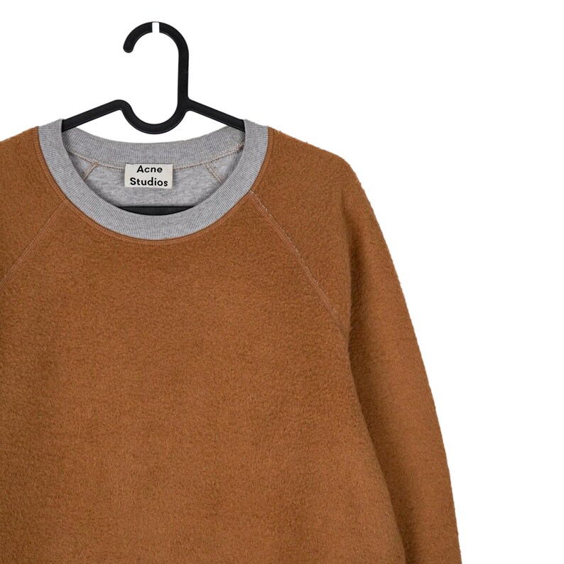 May include: A brown fleece sweatshirt with a grey crew neck and cuffs, hanging on a black hanger. The label "Acne Studios" is visible on the front of the sweatshirt.