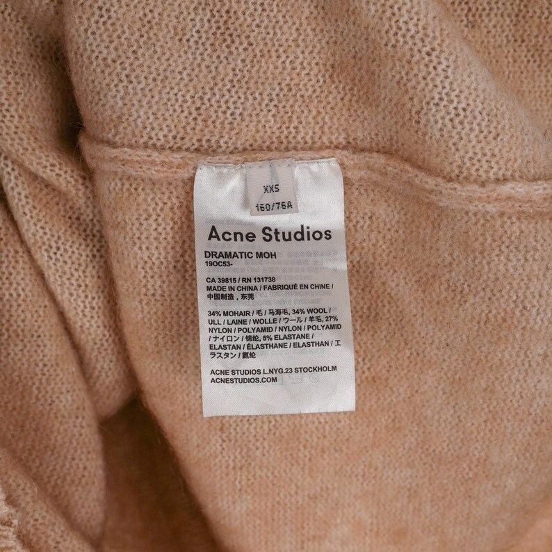 Pu&ograve; includere: Primo piano di un'etichetta di abbigliamento Acne Studios marrone chiaro. L'etichetta mostra la taglia XXS, la composizione del tessuto e il testo "DRAMATIC MOH". Sono visibili anche il marchio e l'indirizzo del sito web.