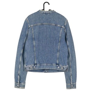 Op de afbeelding: Een lichtblauwe spijkerjas, van achteren gezien, hangend aan een zwarte hanger. De jas heeft een klassiek ontwerp met een kraagloze halslijn, knoopsluitingen en twee borstzakken. De denim heeft een verweerde uitstraling.