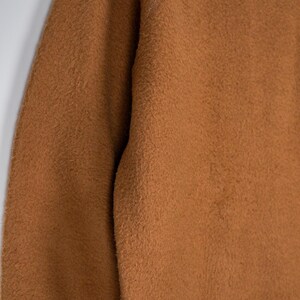 May include: Close-up of a brown, textured fabric. The material has a fuzzy, slightly uneven surface, with a warm, earthy tone. The fabric appears to be thick and possibly knitted or woven, suggesting a cosy garment or textile.