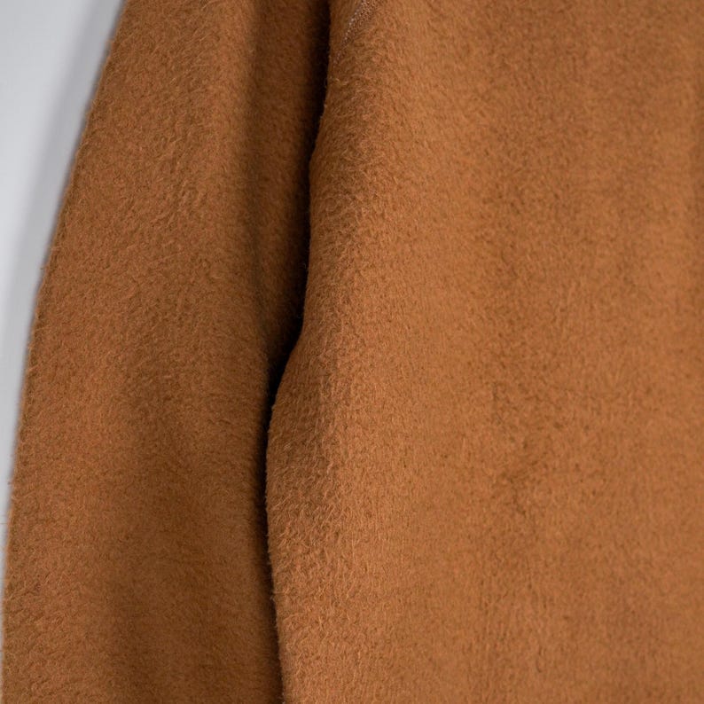 May include: Close-up of a brown, textured fabric. The material has a fuzzy, slightly uneven surface, with a warm, earthy tone. The fabric appears to be thick and possibly knitted or woven, suggesting a cosy garment or textile.