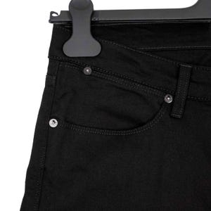 May include: Close-up of black denim jeans hanging on a black hanger. The jeans feature a classic five-pocket design with silver rivets and stitching details. The fabric appears to be a solid, dark black color.