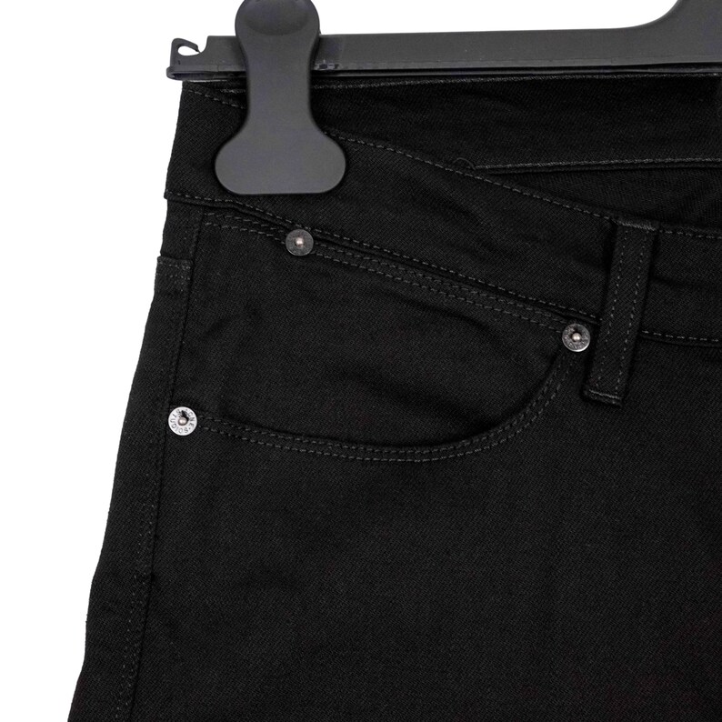 May include: Close-up of black denim jeans hanging on a black hanger. The jeans feature a classic five-pocket design with silver rivets and stitching details. The fabric appears to be a solid, dark black color.