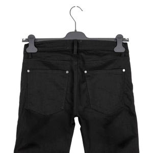 May include: Black denim jeans hanging on a black hanger. The jeans have two back pockets with silver-colored embellishments. The jeans are a solid black color.