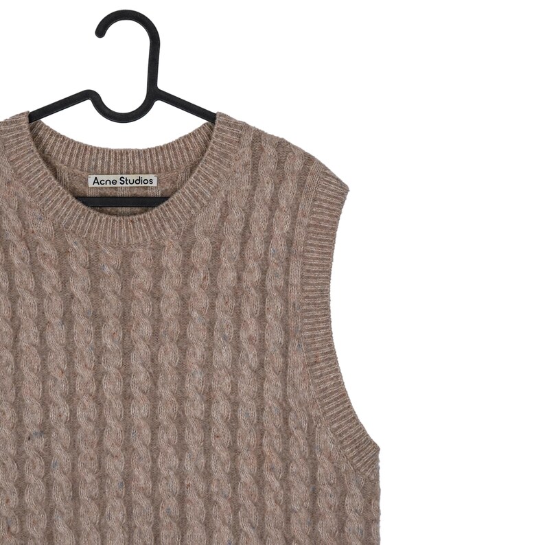 May include: A tan cable-knit sweater vest with a V-neck design. The vest is hanging on a black hanger. The brand name "Acne Studios" is visible on a small tag near the neckline. The vest has a textured, woven appearance.