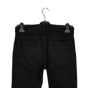 May include: Black denim jeans hanging on a black hanger. The jeans have two back pockets and a classic five-pocket design. The garment is made of a dark, solid-colored fabric.